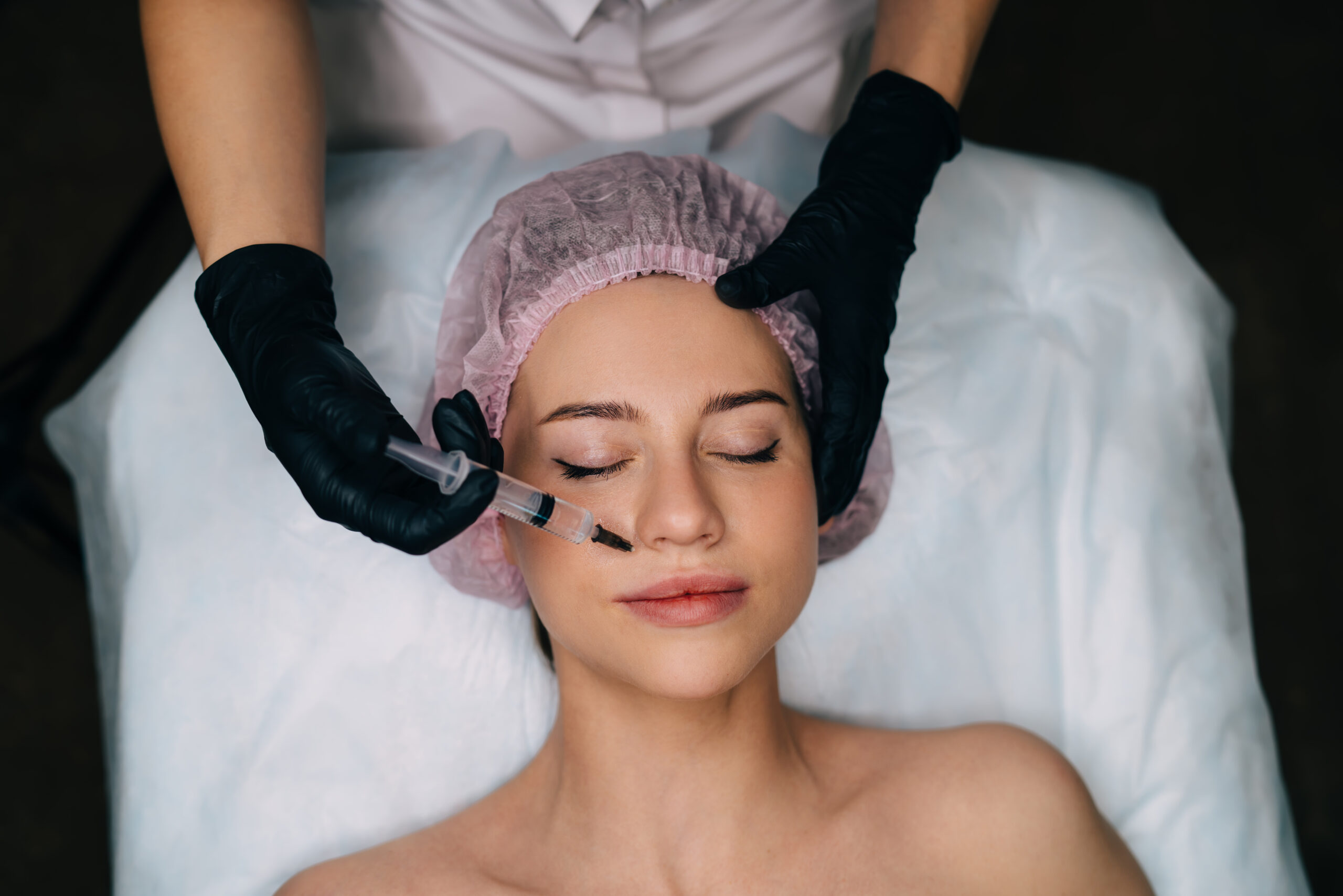 Cropped image of beautician doing botox injection with syringe to girl in beauty salon. Top view of caucasian woman client with closed eyes wear medical cap. Beauty concept. Female master wear gloves